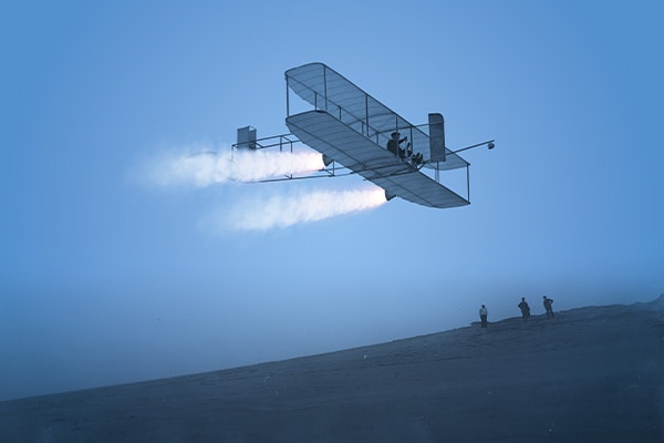 Small biplane flying through the sky with three people watching from a distance